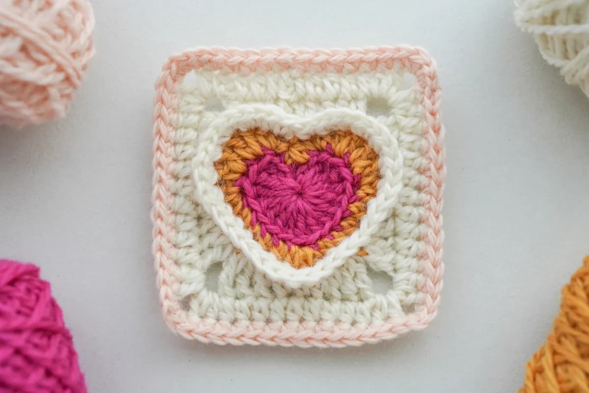 A handmade textured double-heart granny square crocheted in four coordinating colors, featuring a raised heart within a second sculpted heart and a neat square border, photographed directly from above on a soft neutral background with gentle natural daylight, crisp stitch definition, visible front-l