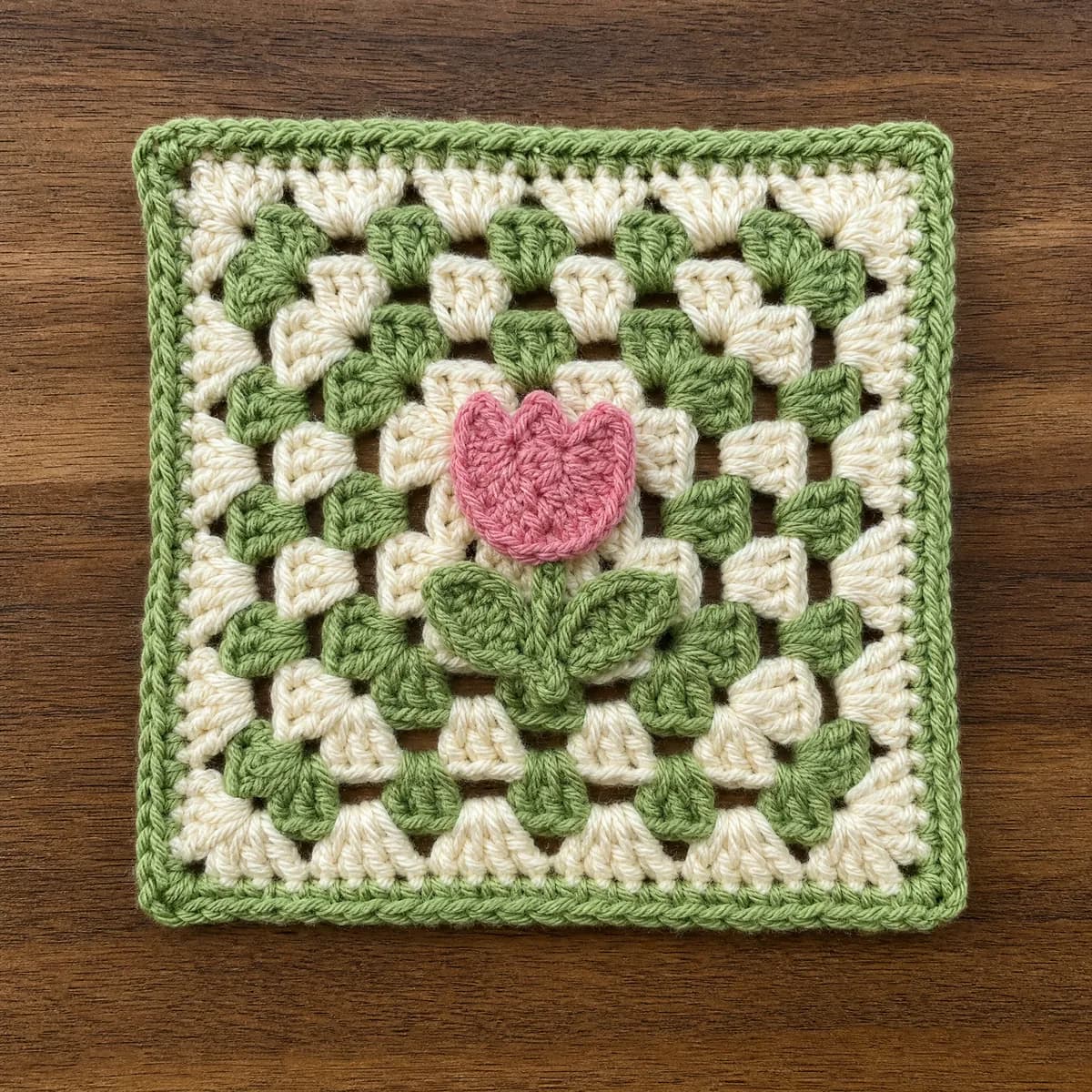 A handmade Little Tulip Granny Square crocheted in cream, leaf green, and soft tulip pink worsted-weight yarn, featuring a classic granny-square background with a raised pink tulip appliqué, slender green stem, and two small green leaves sewn near the center, photographed directly from above on a wa